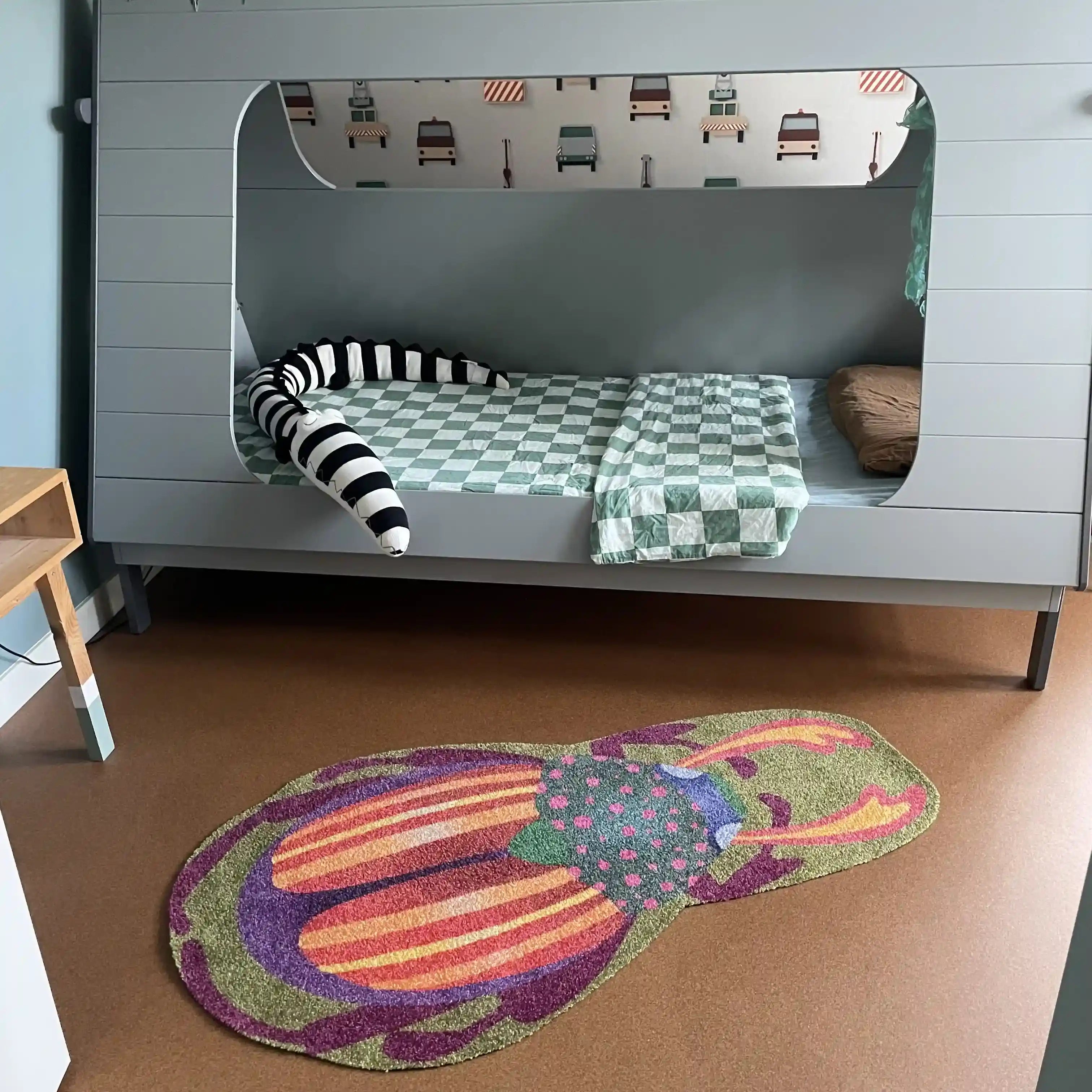 Zacht uitgesneden dierenkleed in de vorm van een kever, gezien van bovenaf, van Studio LilaLine – het kleed toont het volledige lijf van de kever met een ovale vorm en duidelijke segmenten, in mosgroen met accenten van oranje en paars. De pootjes en voelsprieten steken subtiel uit, waardoor de kever herkenbaar en speels oogt. De textuur is zacht en comfortabel, ideaal als fantasierijke speelplek in een kinderkamer.