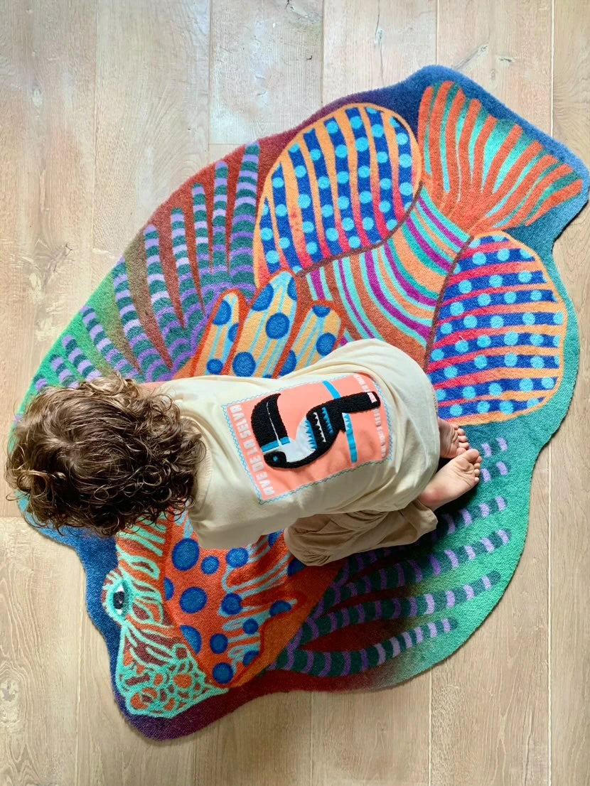 Zacht uitgesneden dierenkleed in de vorm van een koraalduivel van Studio LilaLine – deze fantasierijke vis heeft sierlijke vinnen die wijd uitstaan, in sprekende tinten groen en oranje. De visvorm is dynamisch en decoratief, met speelse strepen en zachte rondingen. Het kleed is heerlijk zacht en nodigt uit tot spelen, liggen of fantaseren in een gezellige hoek van de kinderkamer.