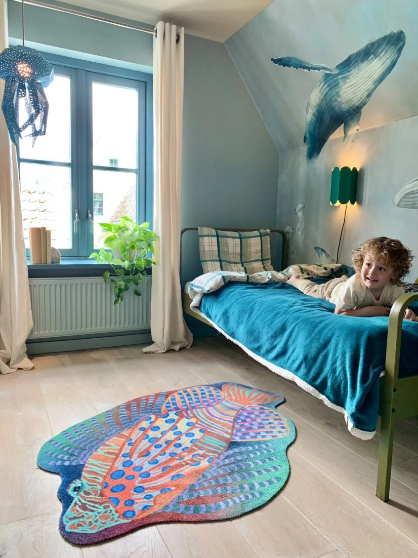 Zacht uitgesneden dierenkleed in de vorm van een koraalduivel van Studio LilaLine – deze fantasierijke vis heeft sierlijke vinnen die wijd uitstaan, in sprekende tinten groen en oranje. De visvorm is dynamisch en decoratief, met speelse strepen en zachte rondingen. Het kleed is heerlijk zacht en nodigt uit tot spelen, liggen of fantaseren in een gezellige hoek van de kinderkamer.