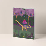 Dubbelgevouwen A6-kaart van Studio LilaLine met op de voorkant een vrolijke dino met een lange nek – waarschijnlijk een brachiosaurus of apatosaurus – die een grote verjaardagstaart op zijn rug draagt. De taart is rijk versierd met kaarsjes en vrolijke kleuren. De illustratie straalt feestelijkheid en zachtheid uit. De binnenkant van de kaart is blanco en klaar voor een persoonlijke boodschap.