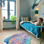 Zacht uitgesneden dierenkleed in de vorm van een koraalduivel van Studio LilaLine – deze fantasierijke vis heeft sierlijke vinnen die wijd uitstaan, in sprekende tinten groen en oranje. De visvorm is dynamisch en decoratief, met speelse strepen en zachte rondingen. Het kleed is heerlijk zacht en nodigt uit tot spelen, liggen of fantaseren in een gezellige hoek van de kinderkamer.