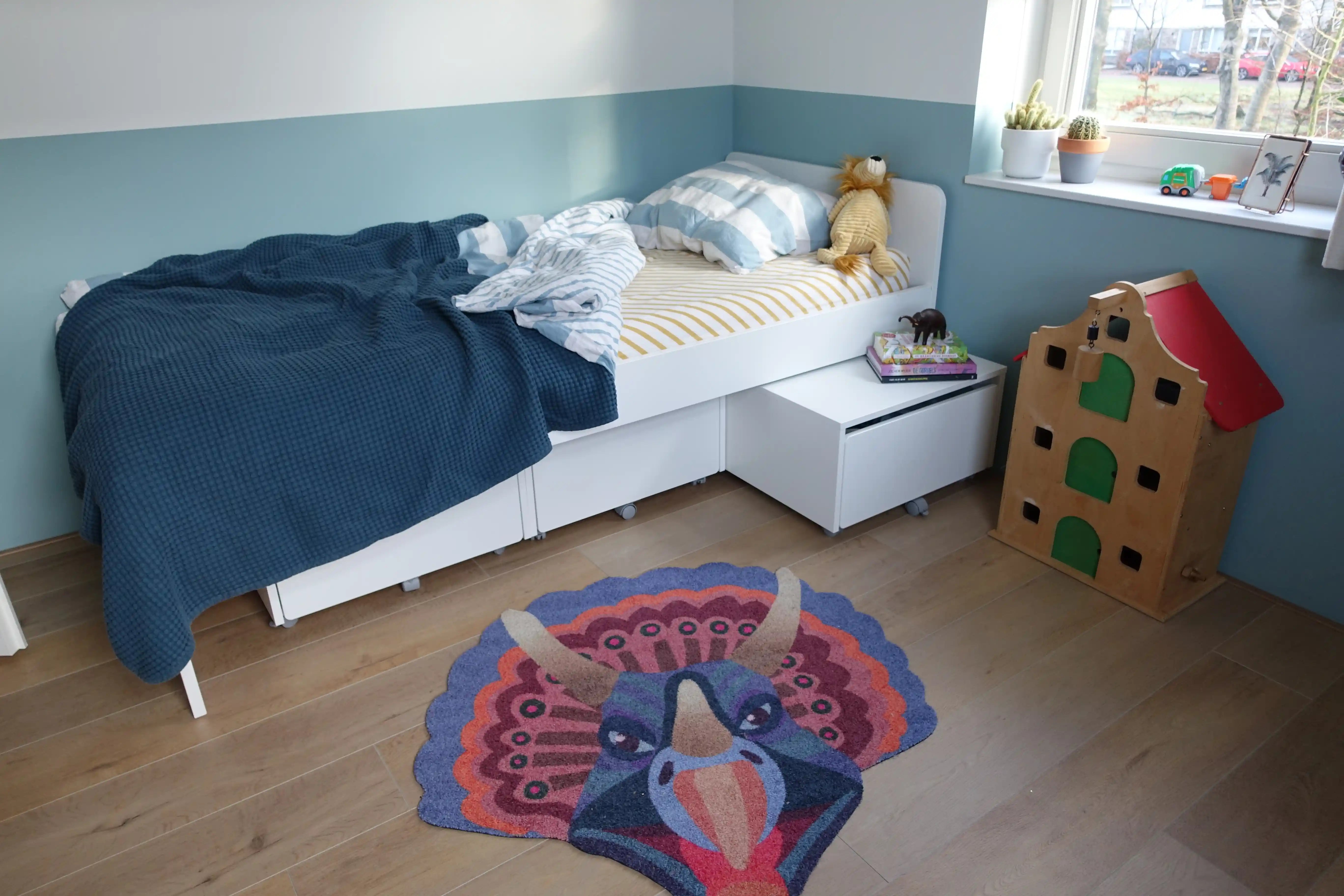 Zacht uitgesneden dierenkleed in de vorm van een triceratopshoofd van Studio LilaLine – dit kleed toont een stoere dinosaurus met drie hoorns en een waaierachtig schild, uitgevoerd in levendige blauw- en oranjetinten. De vorm is krachtig en symmetrisch, met speelse kleurcontrasten die de details extra laten opvallen. Ideaal als zachte speelplek of blikvanger in een kinderkamer met dino-thema.