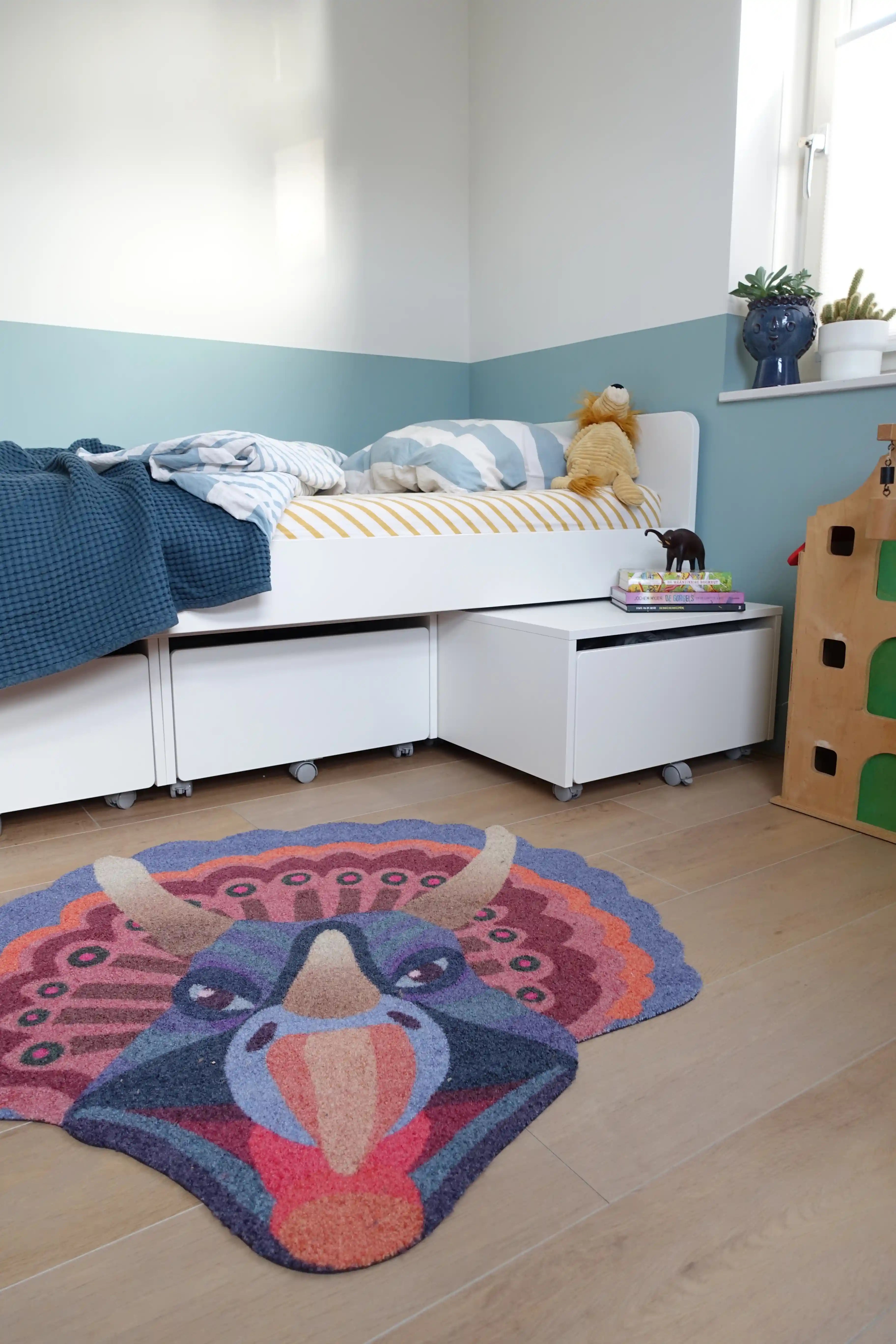 Zacht uitgesneden dierenkleed in de vorm van een triceratopshoofd van Studio LilaLine – dit kleed toont een stoere dinosaurus met drie hoorns en een waaierachtig schild, uitgevoerd in levendige blauw- en oranjetinten. De vorm is krachtig en symmetrisch, met speelse kleurcontrasten die de details extra laten opvallen. Ideaal als zachte speelplek of blikvanger in een kinderkamer met dino-thema.